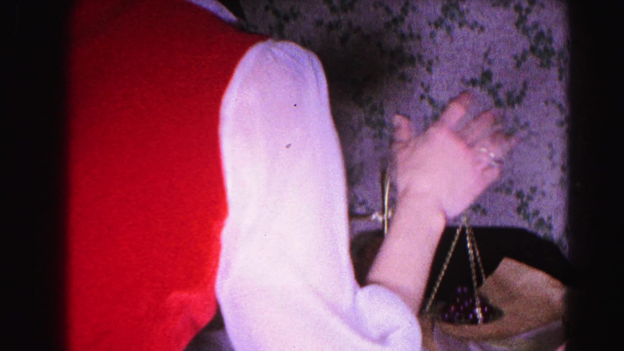 Two women in a domestic interior with floral wallpaper, one in white gesturing over a small brass balance scale.