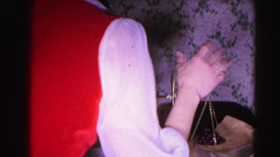 Two women — one in white, one in red — with a brass two-pan balance scale against floral wallpaper; 8mm home movie, early 1960s domestic interior.