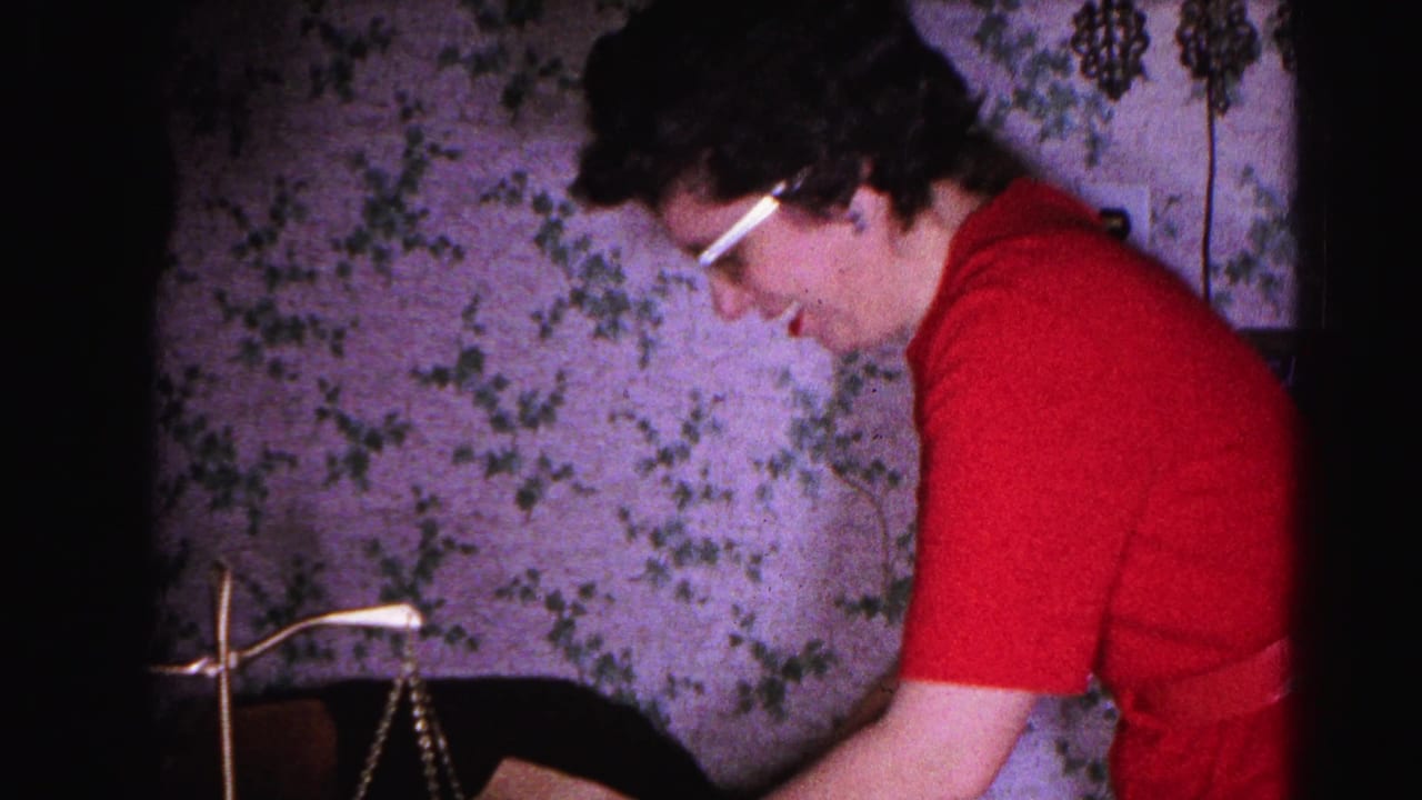 Woman in red short-sleeved top and white-rimmed glasses leaning over a brass balance scale, floral wallpaper behind her.