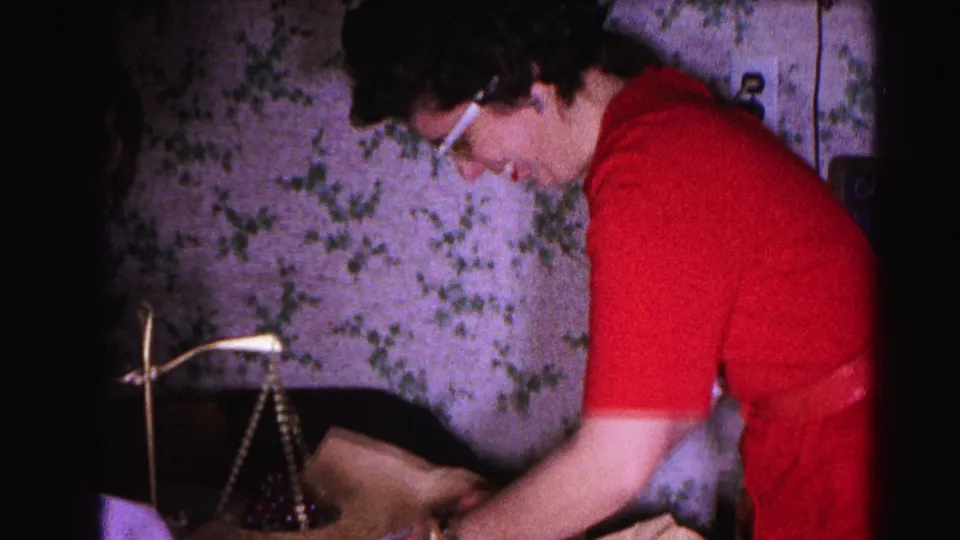 Close view of a woman in red with cat-eye glasses inspecting a brass balance scale, table lamp at left; 1960s domestic interior, 8mm home movie.