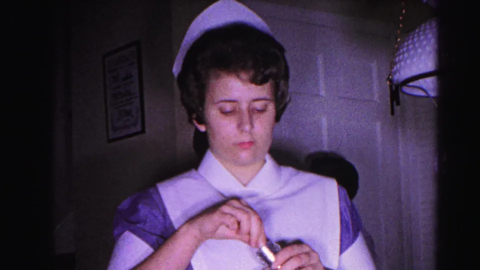 Young woman in 1960s nurse uniform and cap holds a small bottle in a dimly lit domestic interior, likely Hagerstown, Maryland, 1963.