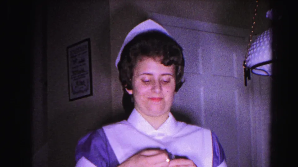 Young woman in 1960s nurse cap and uniform looks downward at a small held object in a domestic interior, circa 1963 Hagerstown, Maryland.