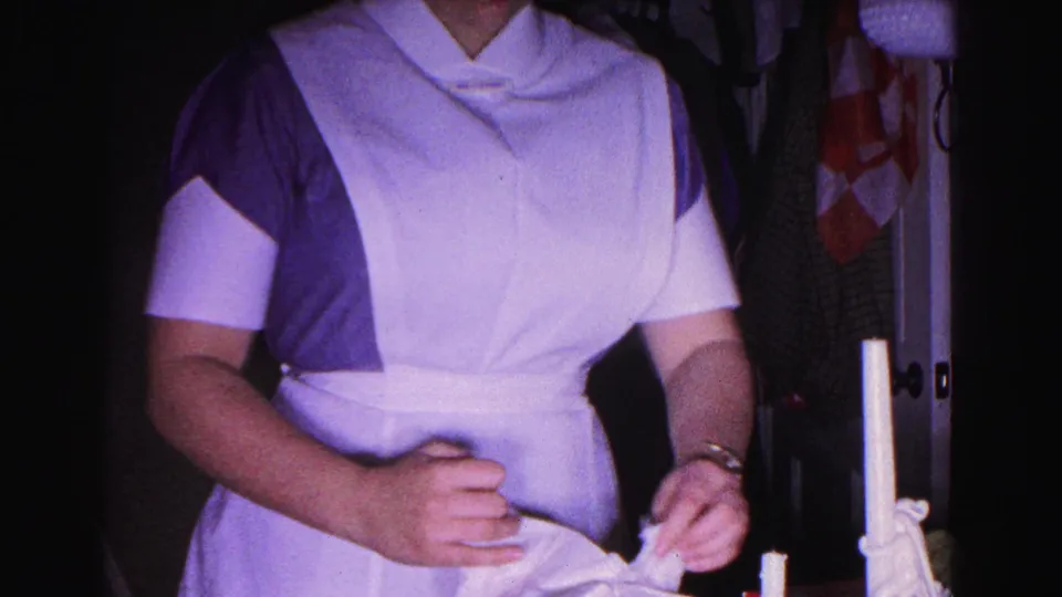 Waist-up shot of a nurse in 1960s-style white bib apron and purple uniform handling white linens; 8mm home movie, Hagerstown, Maryland, 1963.