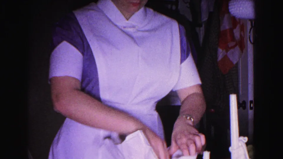 Repeated waist-up view of nurse in 1960s uniform managing white linens in a low-lit interior; 8mm home movie, Hagerstown, Maryland, 1963.