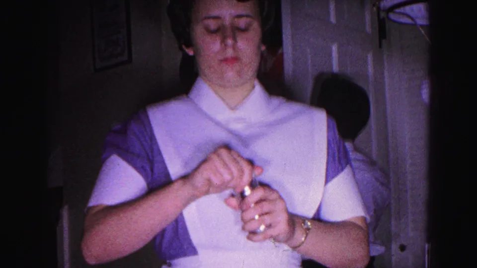Young woman in 1960s nurse uniform with white apron, purple dress, and starched cap handles a small object; face visible; 8mm home movie, Hagerstown, Maryland, 1963.