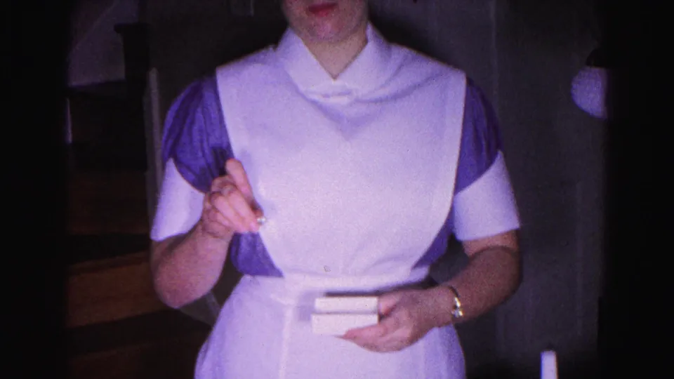 Torso-level close-up of a woman in a 1963-era white nursing uniform with purple shoulder accents, holding small objects in a dim home interior.