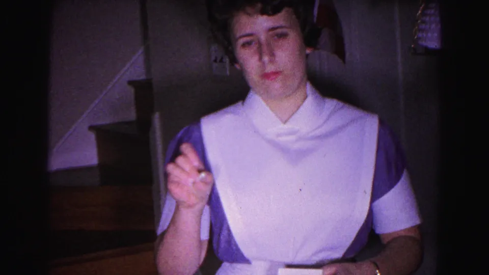 Young woman in 1960s white nursing uniform with purple accents faces camera in a dim home interior; a wooden residential staircase is visible behind her.