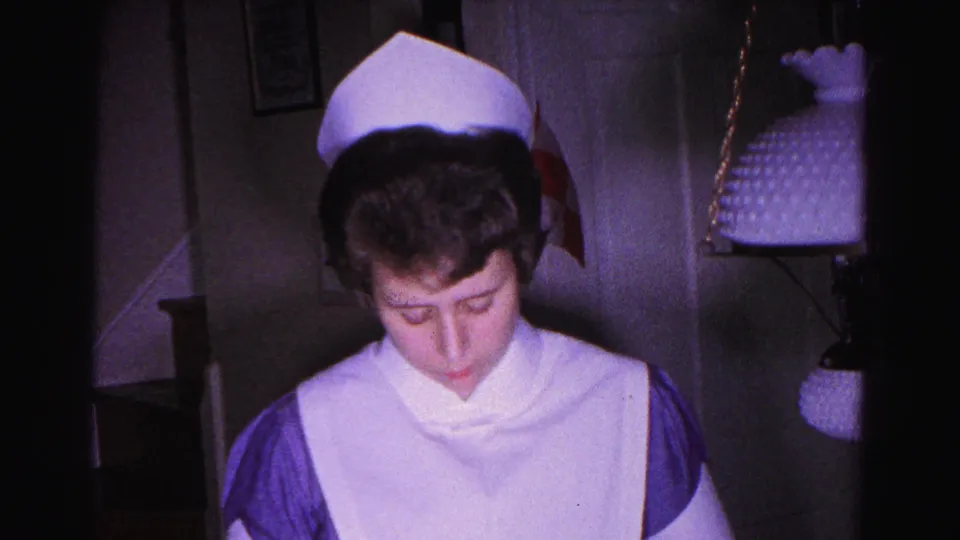 Young woman in traditional white nursing cap and white-and-purple uniform looks down at her hands in a home interior; a hanging pendant lamp on a chain is visible in background.
