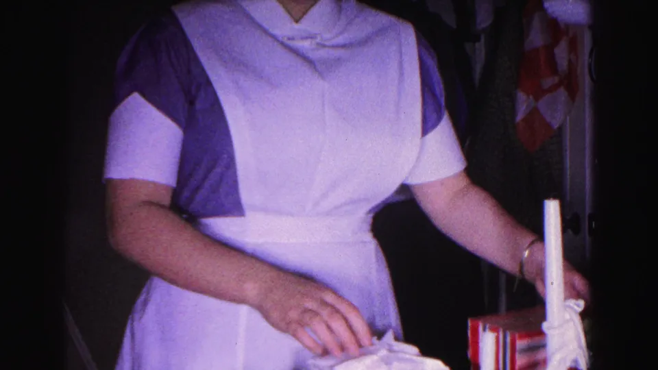 Woman in 1960s nursing uniform handles a white candle and small items at a surface; a red-and-white patterned fabric is visible in the background of the home interior.