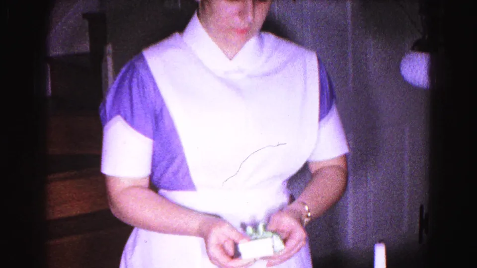1963 8mm home movie frame: woman in blue dress and white apron begins opening a small gift box indoors, Hagerstown, Maryland.