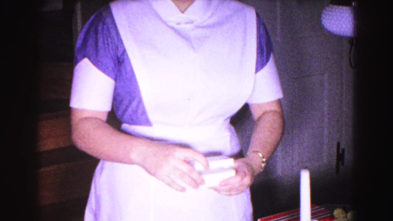Woman in blue dress and white apron holds a flat item from the gift box, white candle and red-white items visible at right.
