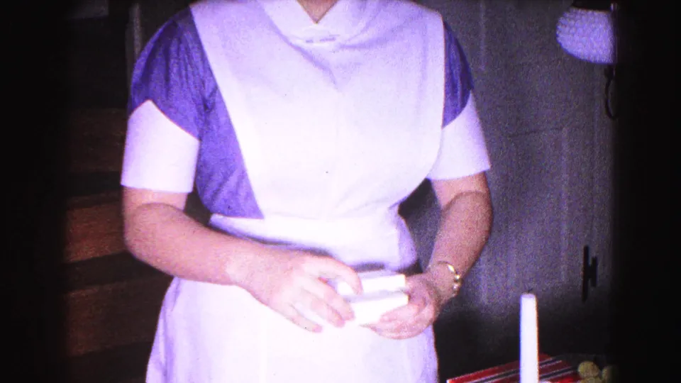 1963 8mm home movie: woman in blue dress and white apron examining gift contents in a domestic holiday setting.