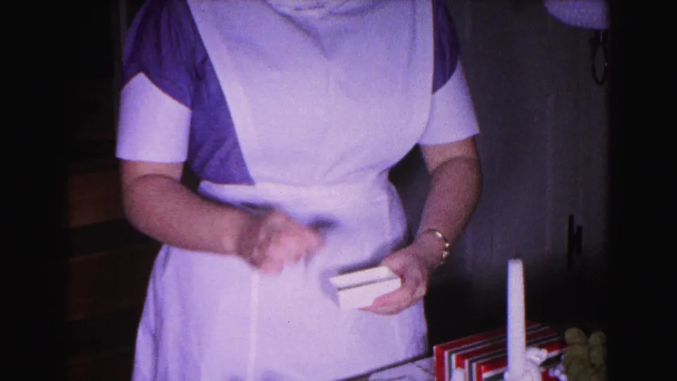 1963 8mm home movie: woman in blue dress and apron actively handling flat gift item in a domestic interior, motion blur on hands.