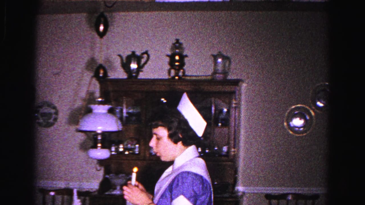 Young woman in nurse's uniform holds a lit candle in a dimly lit dining room; wooden china hutch visible behind her.