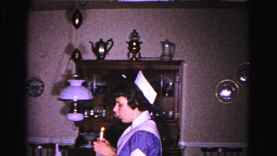 Young woman in 1960s nurse's cap and blue pinafore uniform holds a lit candle in a domestic dining room, Hagerstown, Maryland, circa 1963.