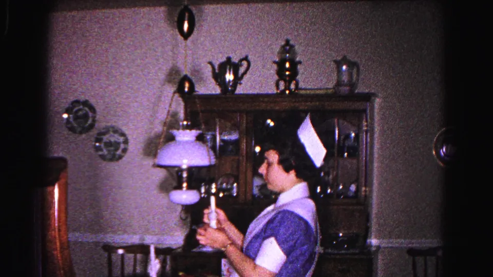 Young woman in 1960s nurse's uniform raises a candle near a hanging lavender-shaded parlor lamp in a domestic dining room, circa 1963.