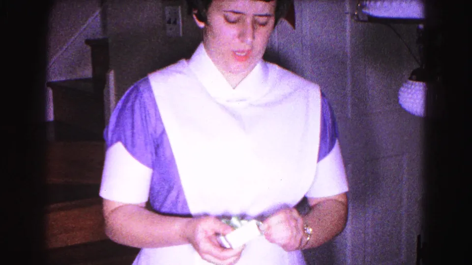 Close view of young woman in blue 1960s nurse's uniform and white cap looking down at candle, with staircase railing and partial hanging lamp behind her, circa 1963.