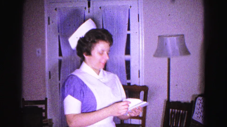 A woman in a 1960s-era blue-and-white nurse uniform and starched cap reads a paper in a domestic interior, Hagerstown, Maryland, circa 1963.