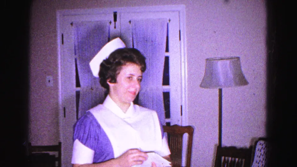 A woman in a 1960s nurse uniform smiles while looking up from a letter in a domestic living room, Hagerstown, Maryland, circa 1963.