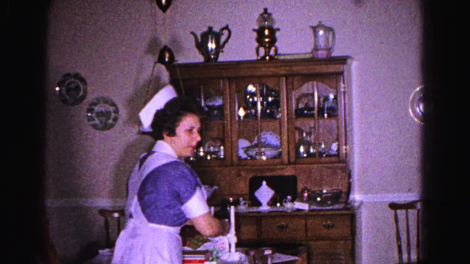 A woman in a 1960s nurse uniform stands beside a wooden china hutch with a silver tea service in a domestic dining room, Hagerstown, Maryland, circa 1963.