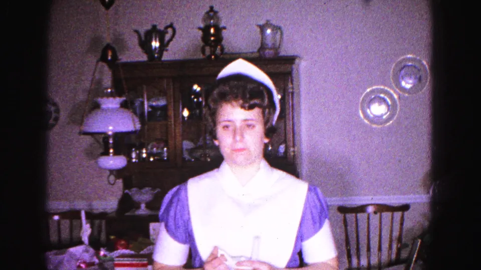 A woman in a 1960s nurse uniform looks directly into the camera in a domestic dining room with a wooden hutch and table lamp behind her, Hagerstown, Maryland, circa 1963.