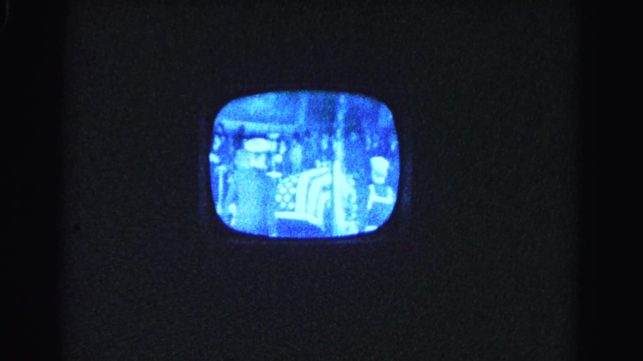 Darkened room with a vintage CRT television, its blue-lit screen displaying a grainy monochrome broadcast with indistinct figures.