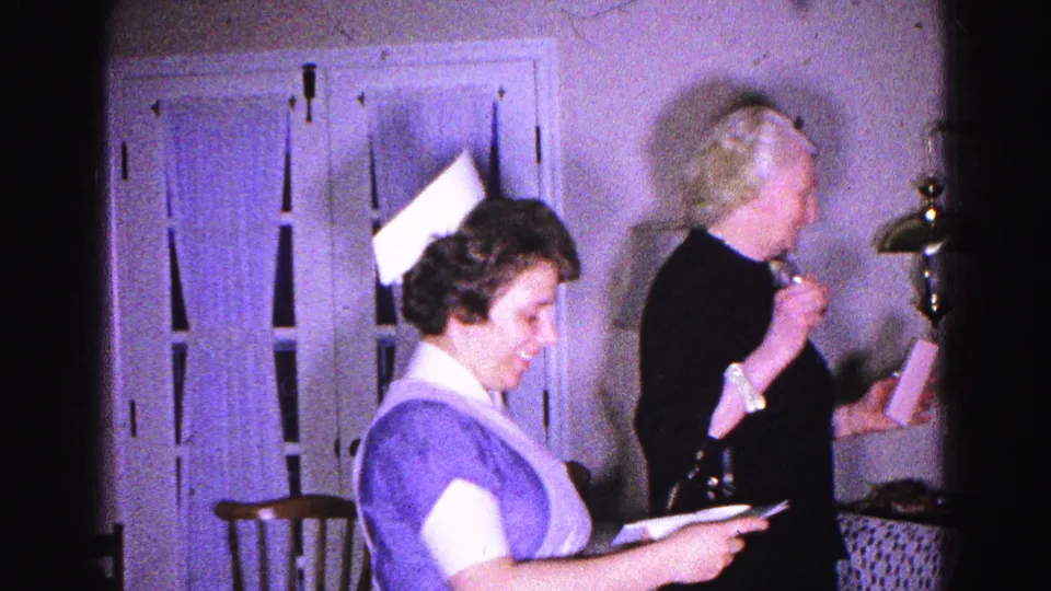 8mm home-movie frame of a smiling nurse in white cap and purple uniform holding a letter, with an older woman behind her in a 1963 home interior.
