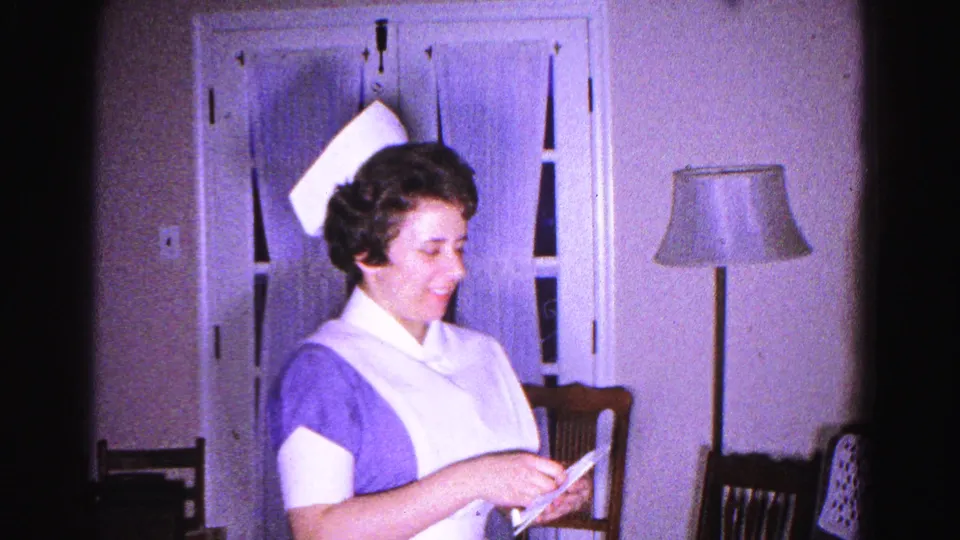 8mm home-movie close shot of a smiling nurse in white cap and purple uniform reading a letter in a 1960s home interior, Hagerstown, Maryland.