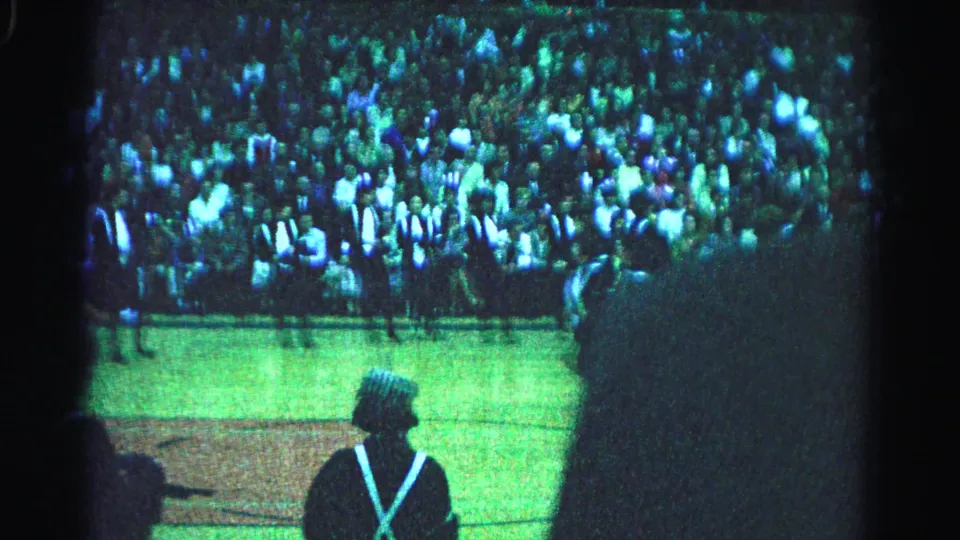1963 Hagerstown, Maryland: uniformed marchers with white X-cross-straps on green field; large crowd fills background stands; cameraman shadow in foreground.