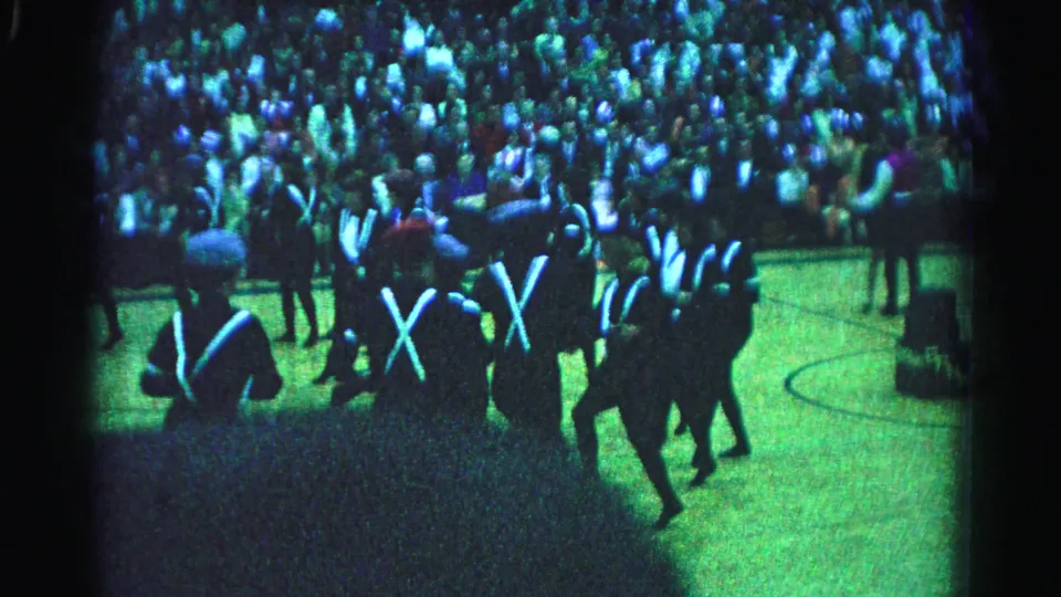 1963 8mm home-movie footage from Hagerstown, Maryland: marching band members in dark uniforms with white cross straps on a green surface, large spectator crowd behind under blue-green stage lighting.