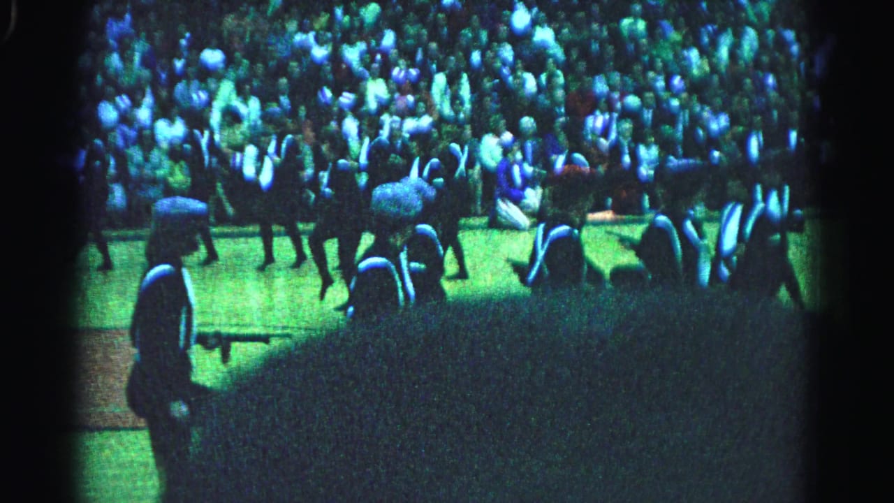 8mm home movie circa 1963 from Hagerstown, Maryland: a single marching band member in dark uniform with white cross straps stands in the foreground, crowd visible behind under blue-green artificial lighting.