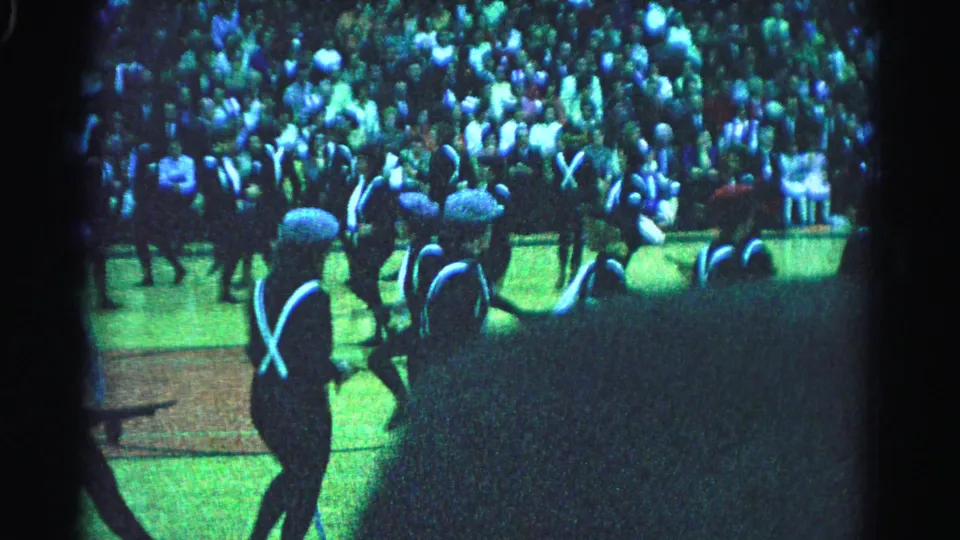 Mid-wide 8mm home-movie frame from a 1963 marching band event in Hagerstown, Maryland, showing multiple uniformed performers in formation under artificial blue-green lighting with a dense crowd behind.