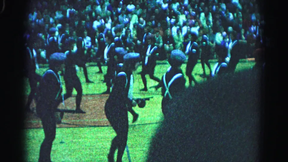 Closing frame of 1963 8mm footage from Hagerstown, Maryland, showing marching band members with white cross-strap uniforms on a green performance surface with a large public audience visible in background.