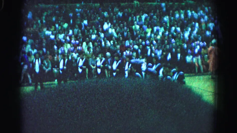 Mid-clip 8mm frame of large outdoor gathering in Hagerstown Maryland 1963, showing crowd rows with prominent cast shadow in foreground.