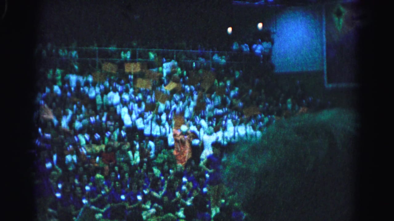 Large crowd in stadium bleachers at night under blue-green artificial lighting, 8mm footage circa 1963.