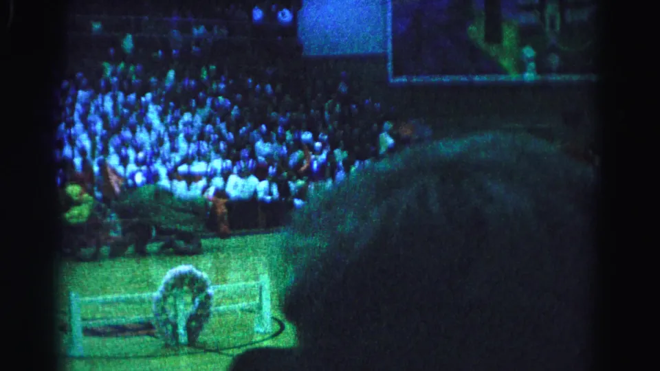 Frame from a 1963 Hagerstown indoor performance showing a horse costume performer near the barrier, large audience in bleachers, stage lighting casting blue-green tones.