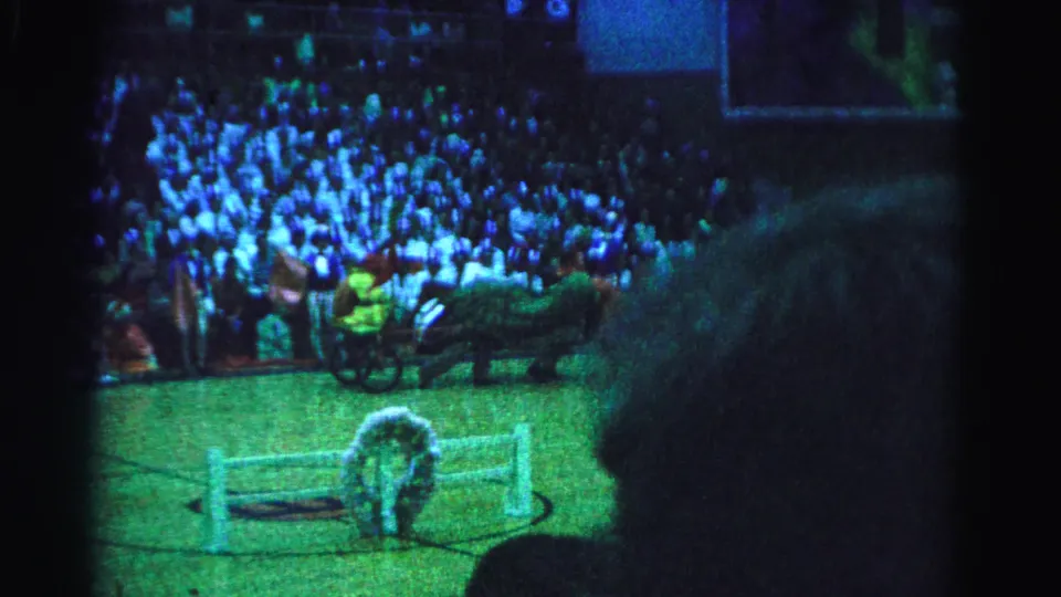 Final frame from a 1963 Hagerstown gymnasium performance showing a chariot with rider alongside horse-costumed performers, crowd filling the bleachers under blue-green lighting.
