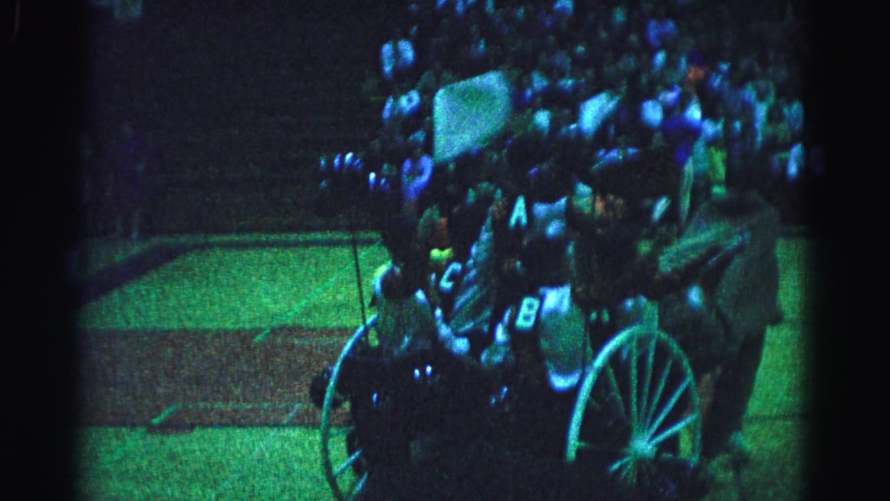 Horse-drawn chariot with large spoked wheels moves through a venue, riders in lettered costumes visible, dense crowd in background under blue-green stage lighting.