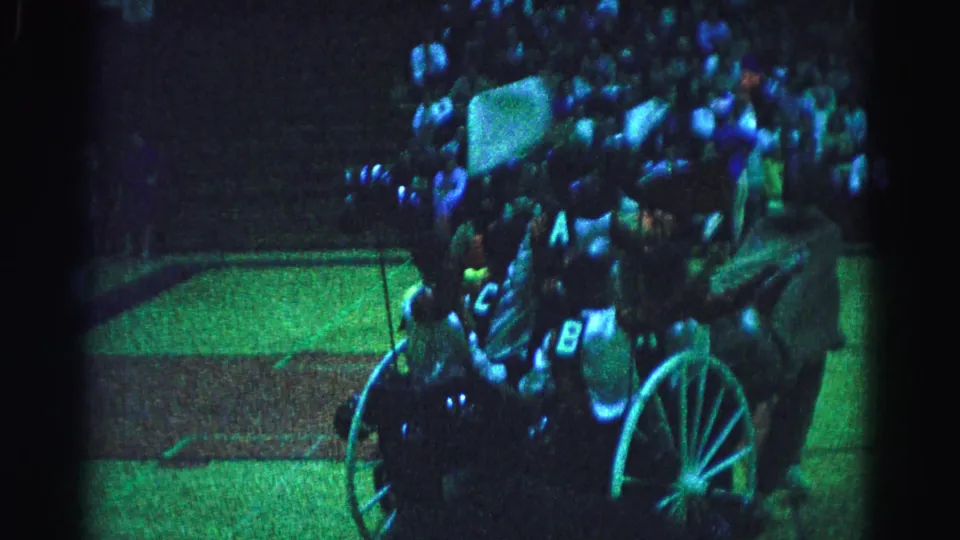 8mm home movie frame, possibly 1963 Hagerstown Maryland, showing a horse-drawn chariot with spoked wheels, riders with letter-marked costumes, and a packed crowd under blue-green stage lighting.