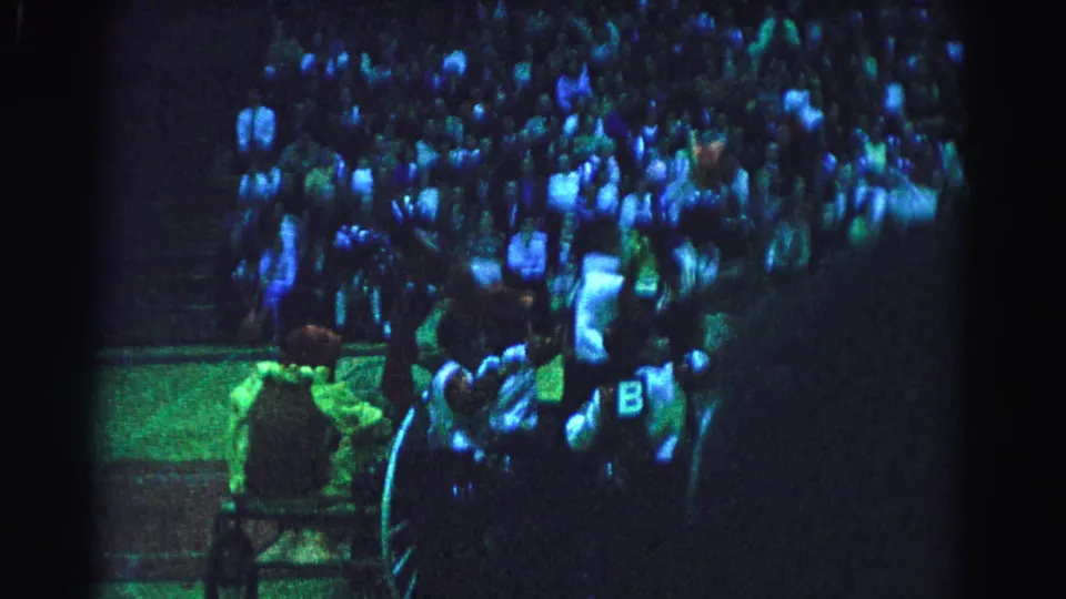 8mm frame showing chariot wheel and costumed rider with letter B insignia, dense audience in background, strong blue-green stage lighting, possibly a 1963 Hagerstown Maryland arena event.
