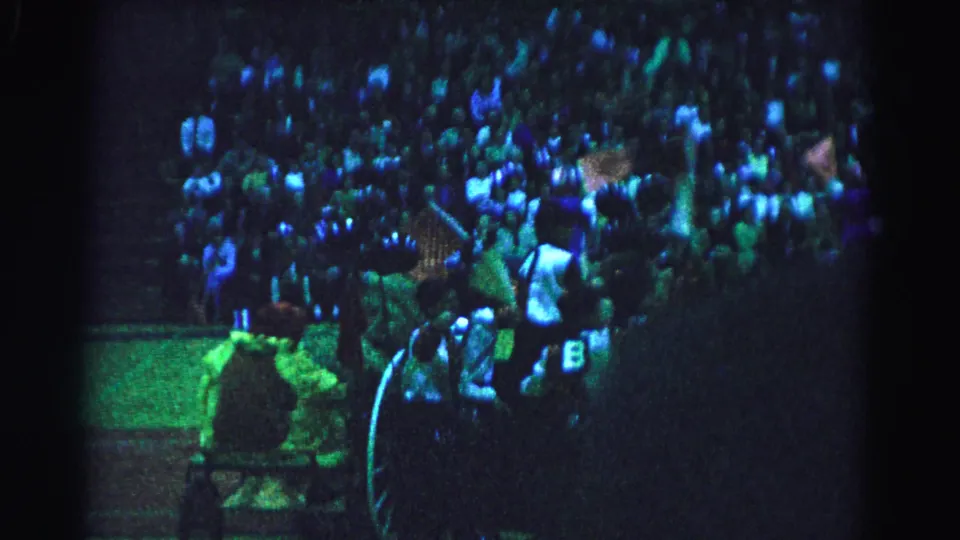 8mm frame showing wide bleacher audience and performers with chariot in the lower foreground, green-blue stage lighting, possibly a 1963 indoor arena event in Hagerstown Maryland.
