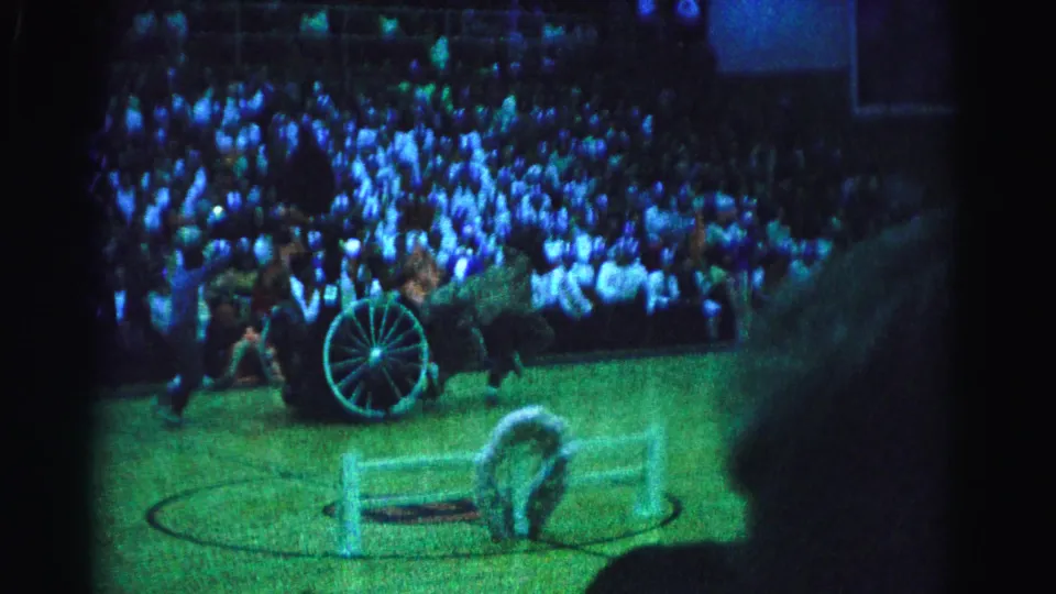 8mm frame showing full arena floor and multi-level bleacher crowd; horse and chariot visible at lower right, possible center-floor circle marking below, blue-green stage lighting, circa 1963.