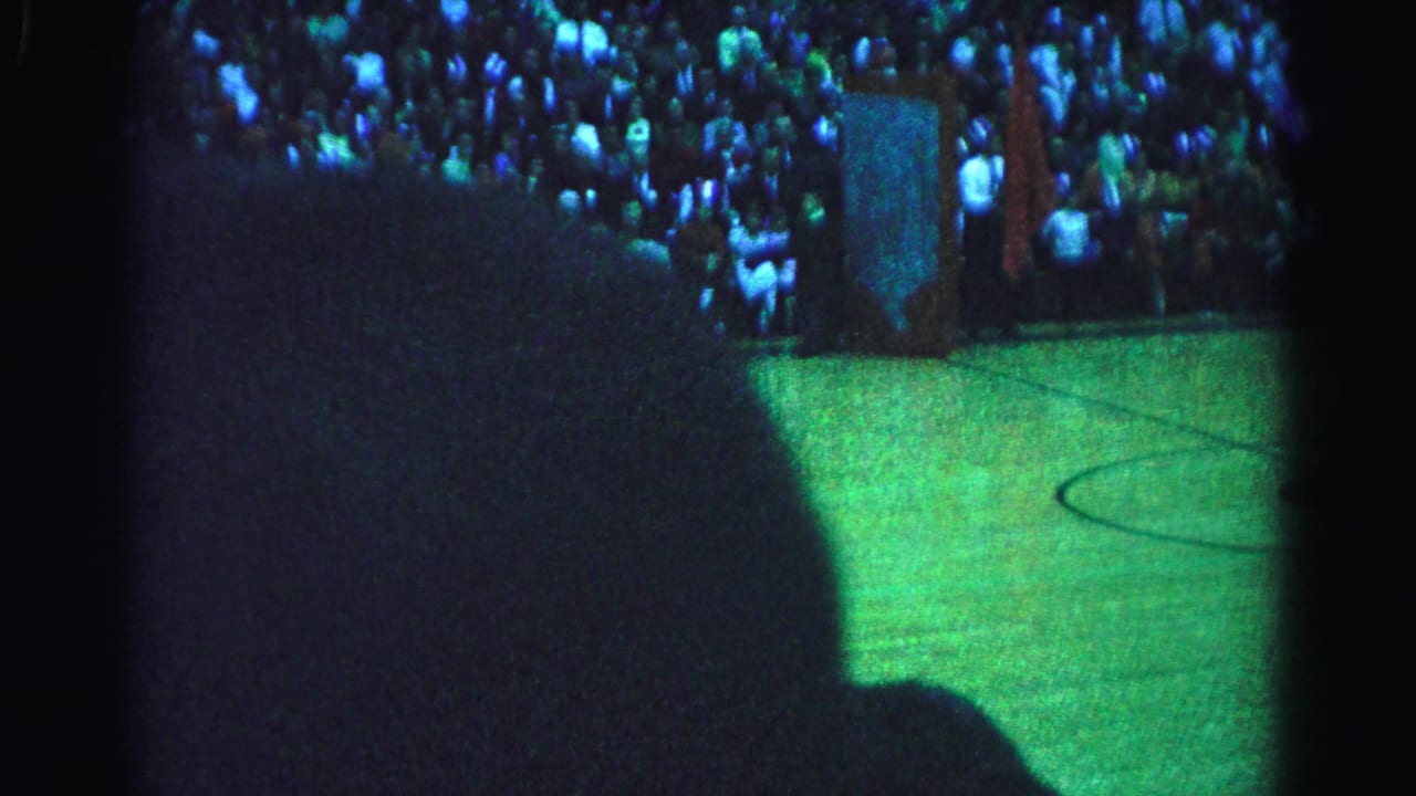 Dark gymnasium interior with large audience filling bleachers; a viewer's foreground silhouette partially obscures the lower frame.