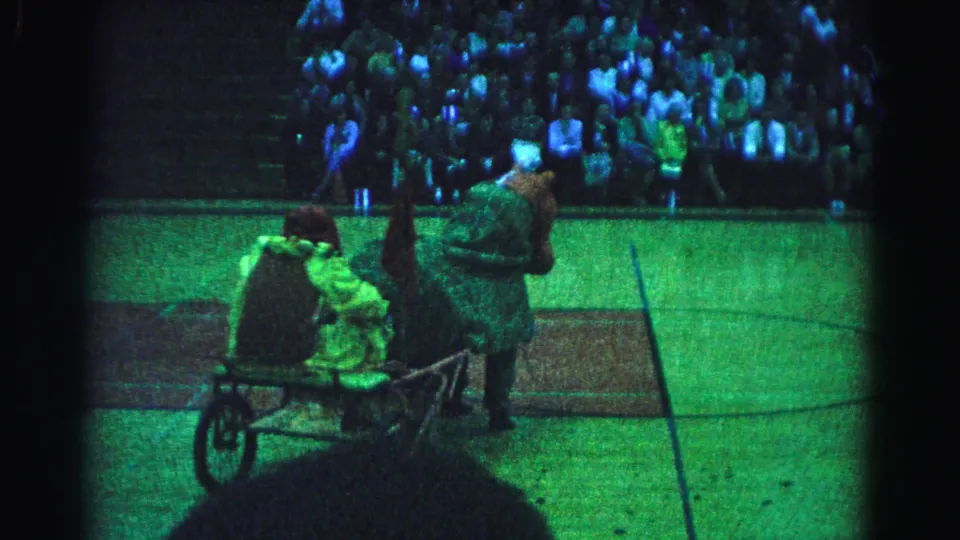 8mm frame from a 1963 Hagerstown, Maryland school reenactment; a costumed performer pushes a wheeled cart carrying a second figure across the green-tinted gymnasium floor as the bleacher crowd watches.