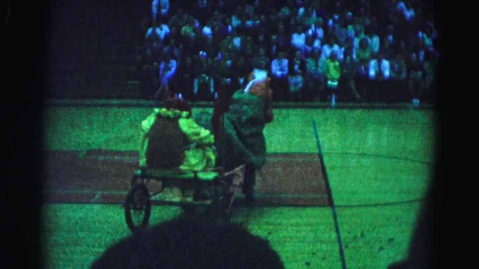 8mm home movie frame from a 1963 Hagerstown, Maryland school reenactment; two costumed performers with a wheeled cart occupy the center of a basketball court, with a large bleacher audience behind them.