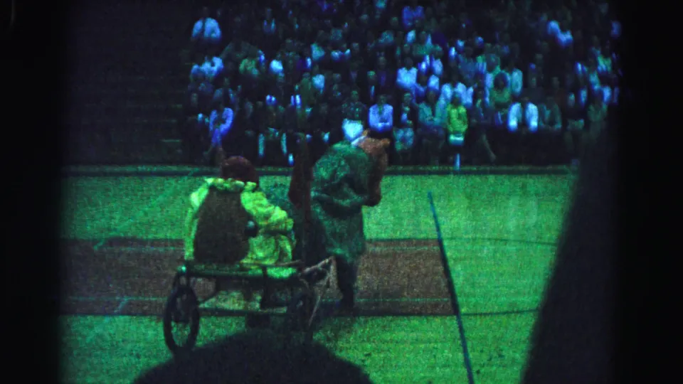 Final 8mm frame from a 1963 Hagerstown, Maryland school play; a costumed performer wheels a cart with a passenger across the court floor as the packed bleacher crowd watches under dim gymnasium lighting.