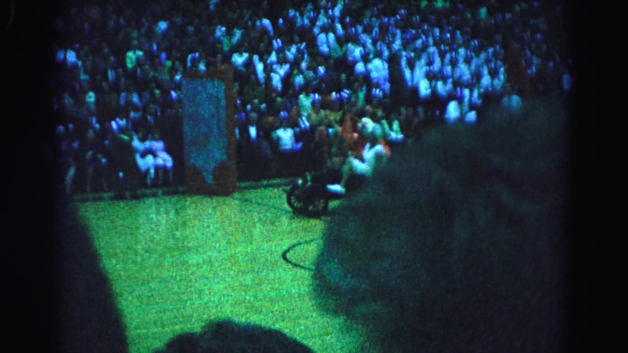 Two costumed performers riding a small vehicle across a green basketball court at what appears to be a 1963 Hagerstown, Maryland arena event, shot on 8mm from the bleachers.