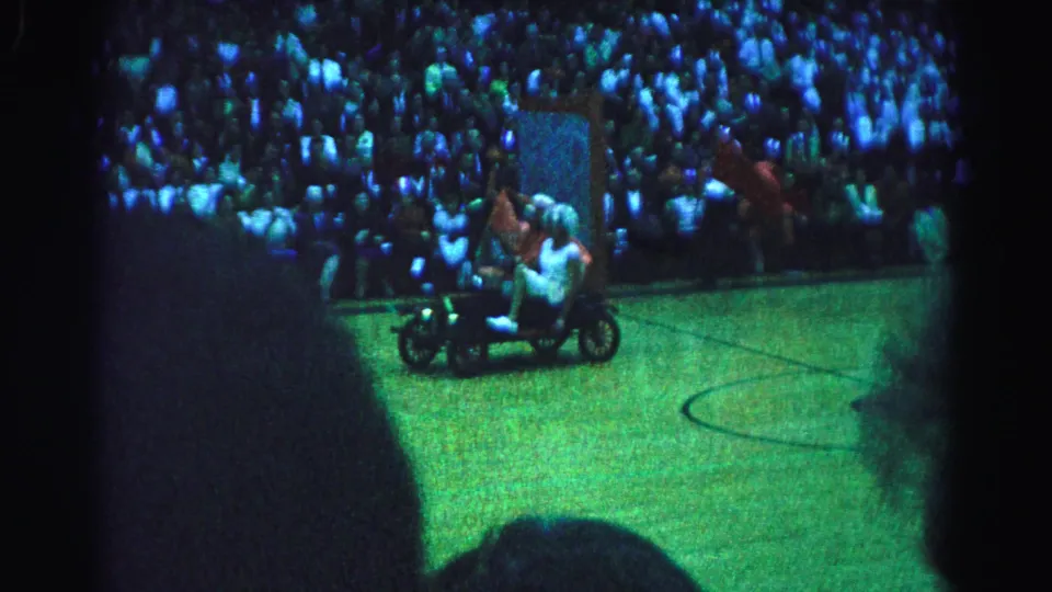Two costumed figures riding a small vehicle across an indoor basketball court floor, filmed from the bleachers at what appears to be a 1963 event in Hagerstown, Maryland.