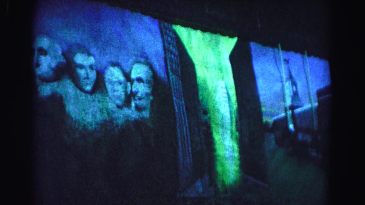 8mm film frame showing two to three presidential faces in a Mount Rushmore projection display after a slight rightward pan, blue-green illumination, circa 1963.