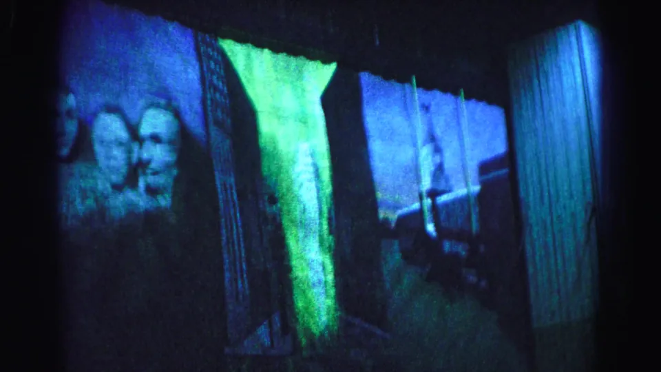 8mm frame with camera panned further right: two Mount Rushmore faces at left edge, a vertical architectural column or pillar emerging at center, blue-green colored lighting, circa 1963.